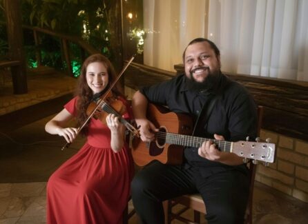 Duo musical com violinista de vestido vermelho e guitarrista barbudo sorrindo, tocando em evento intimista.