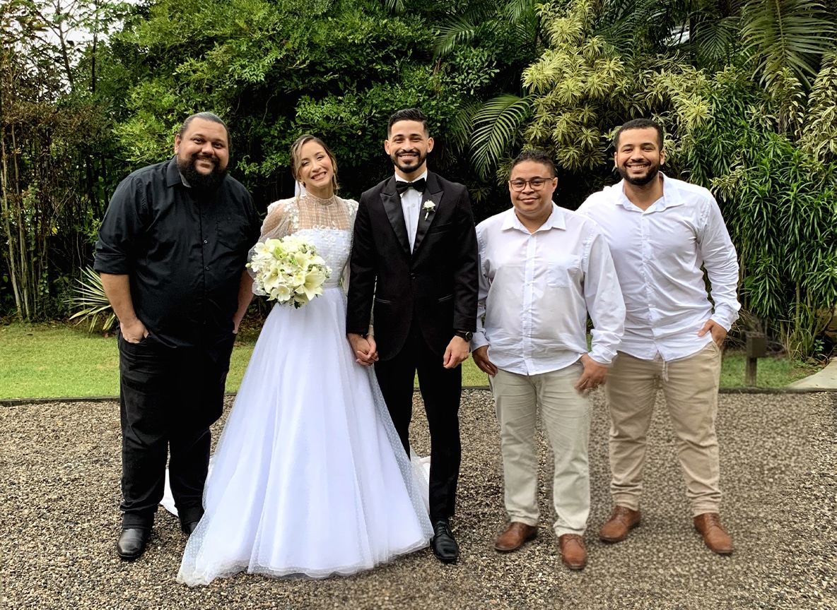 Casal de noivos sorridente posando com três músicos da banda de casamento em cenário verde ao ar livre.