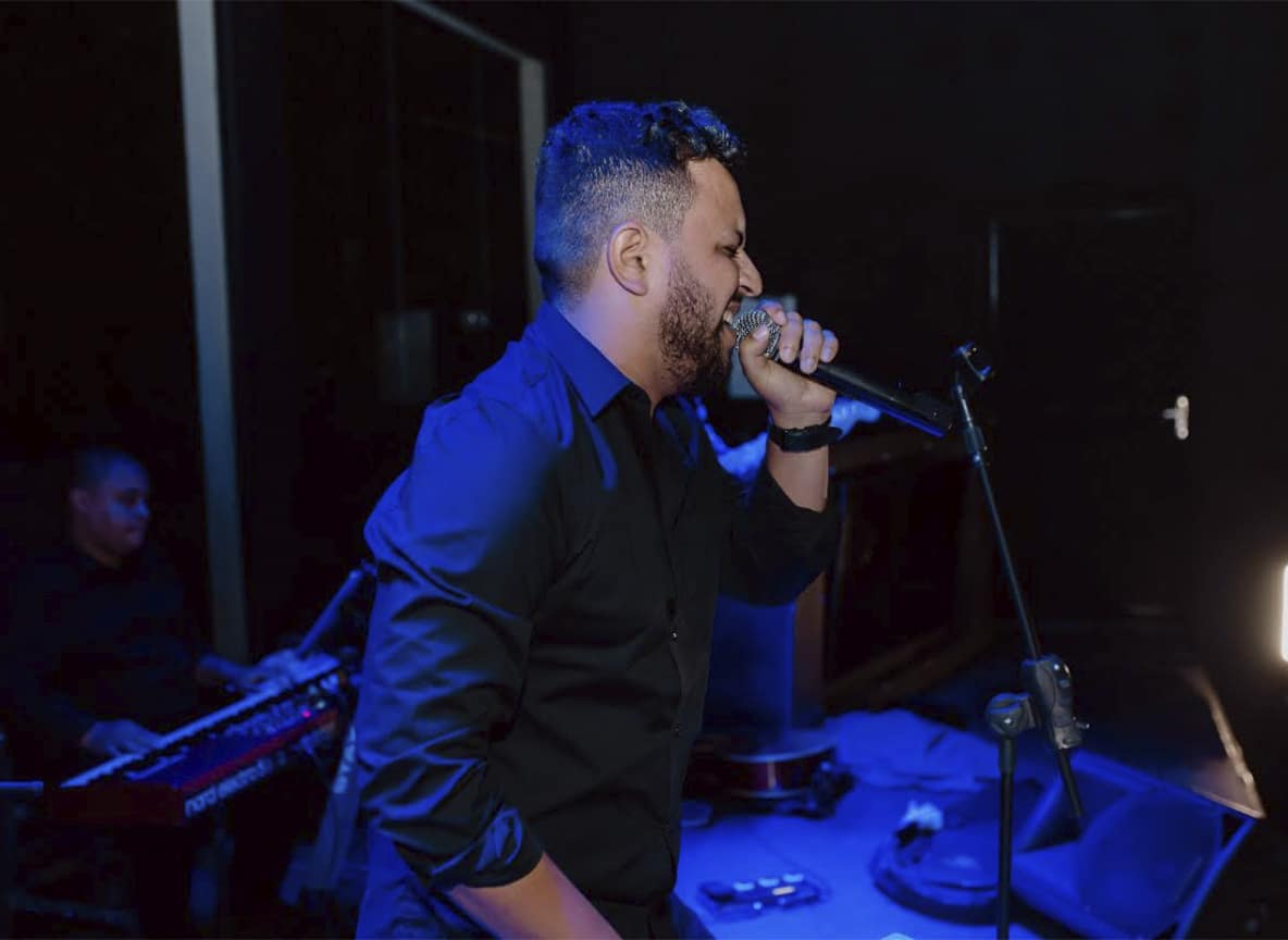 Cantor com camisa social escura no microfone em show noturno com luz azul e tecladista ao fundo.