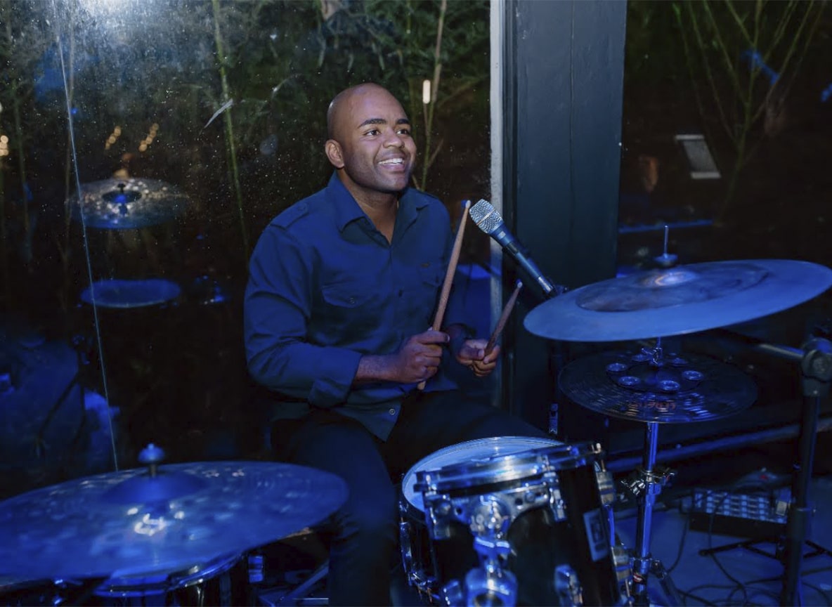 Baterista sorridente tocando bateria em evento noturno com iluminação azul.