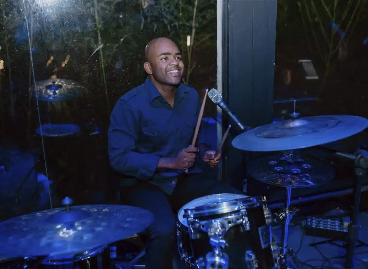 Baterista sorridente tocando bateria em evento noturno com iluminação azul.