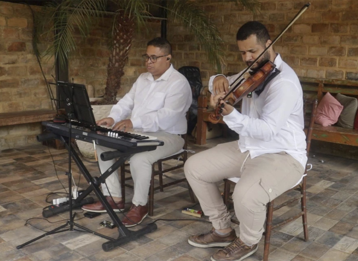 Dupla musical com violinista e tecladista tocando em evento ao ar livre com decoração rústica.