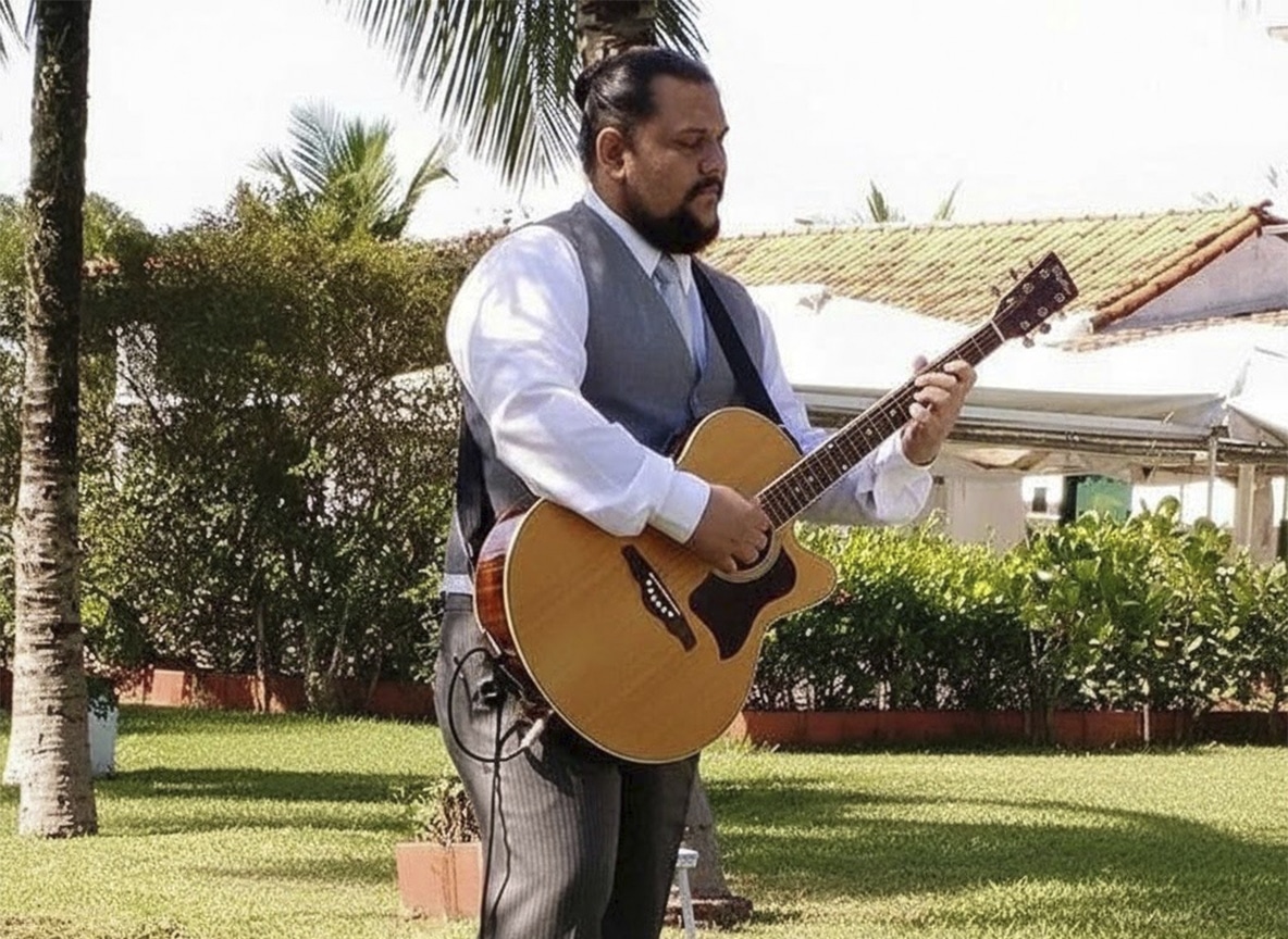 Músico barbudo toca violão em performance solo ao ar livre em evento diurno.
