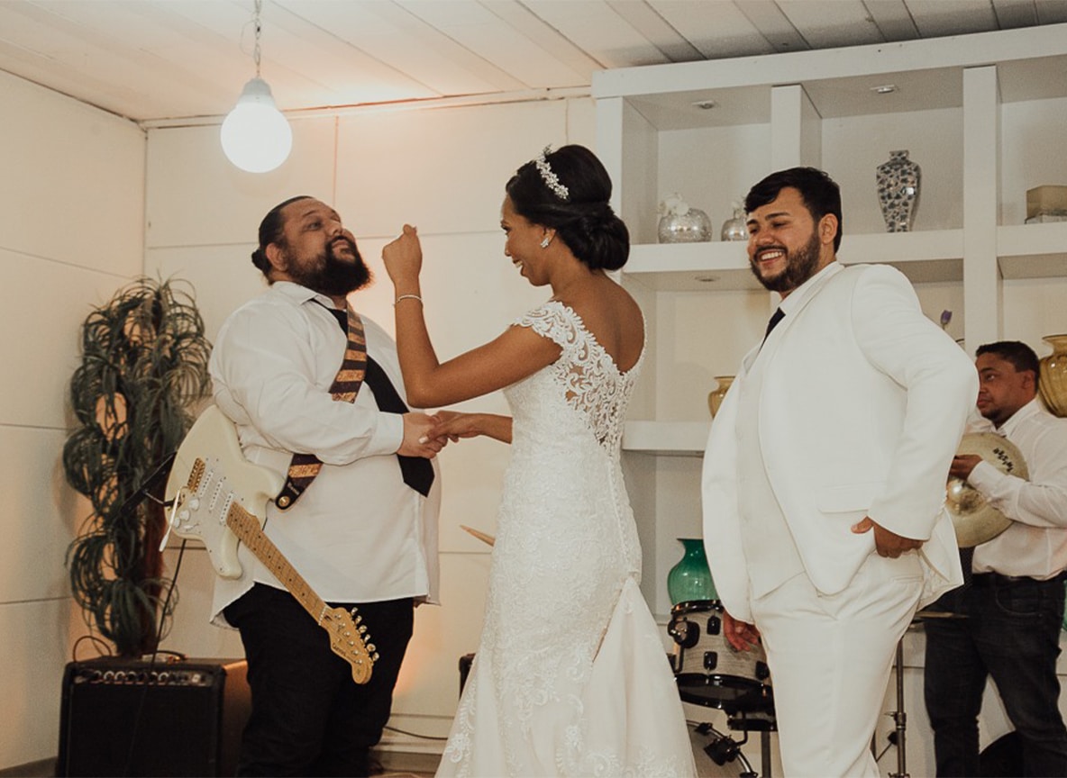 Casal de noivos em vestido branco de renda e terno branco interagindo com músico/cantor que toca guitarra durante festa de casamento com banda.